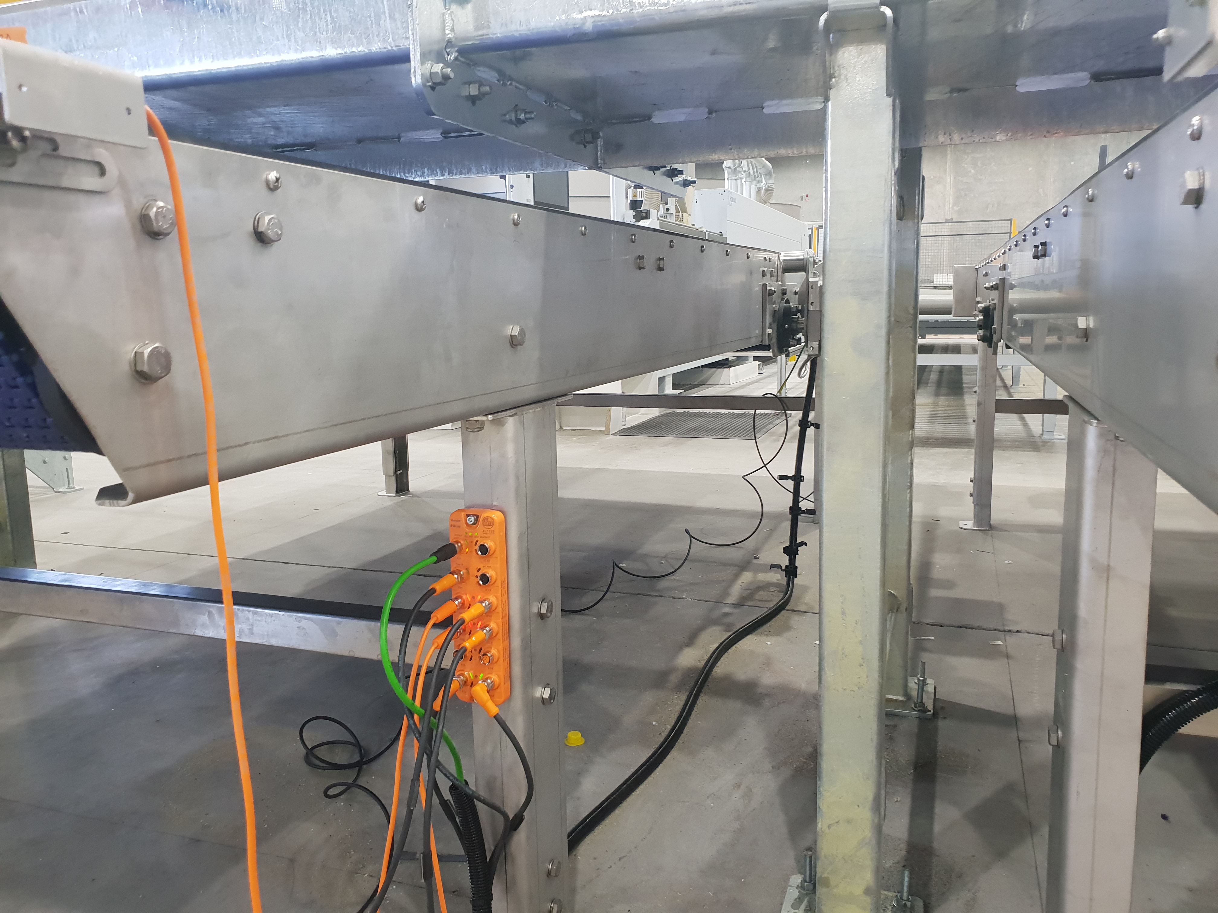 Low-angle view of a stainless conveyor framework with an IFM IO-Link master block and orange / black M12 cabling, before commissioning