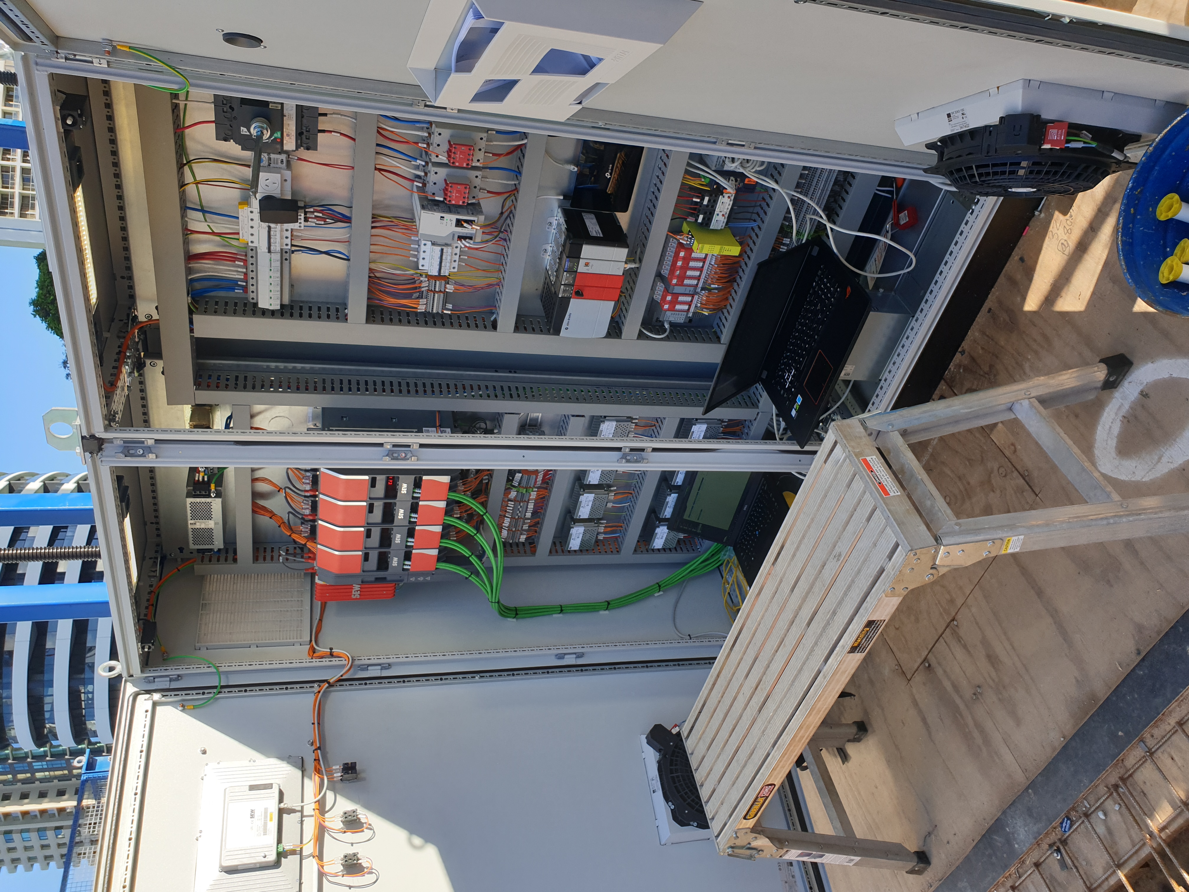 Rooftop MCC commissioning — open switchboards with SEW VFDs and Allen-Bradley CompactLogix, laptops on a hop-up, Gold Coast tower block backdrop